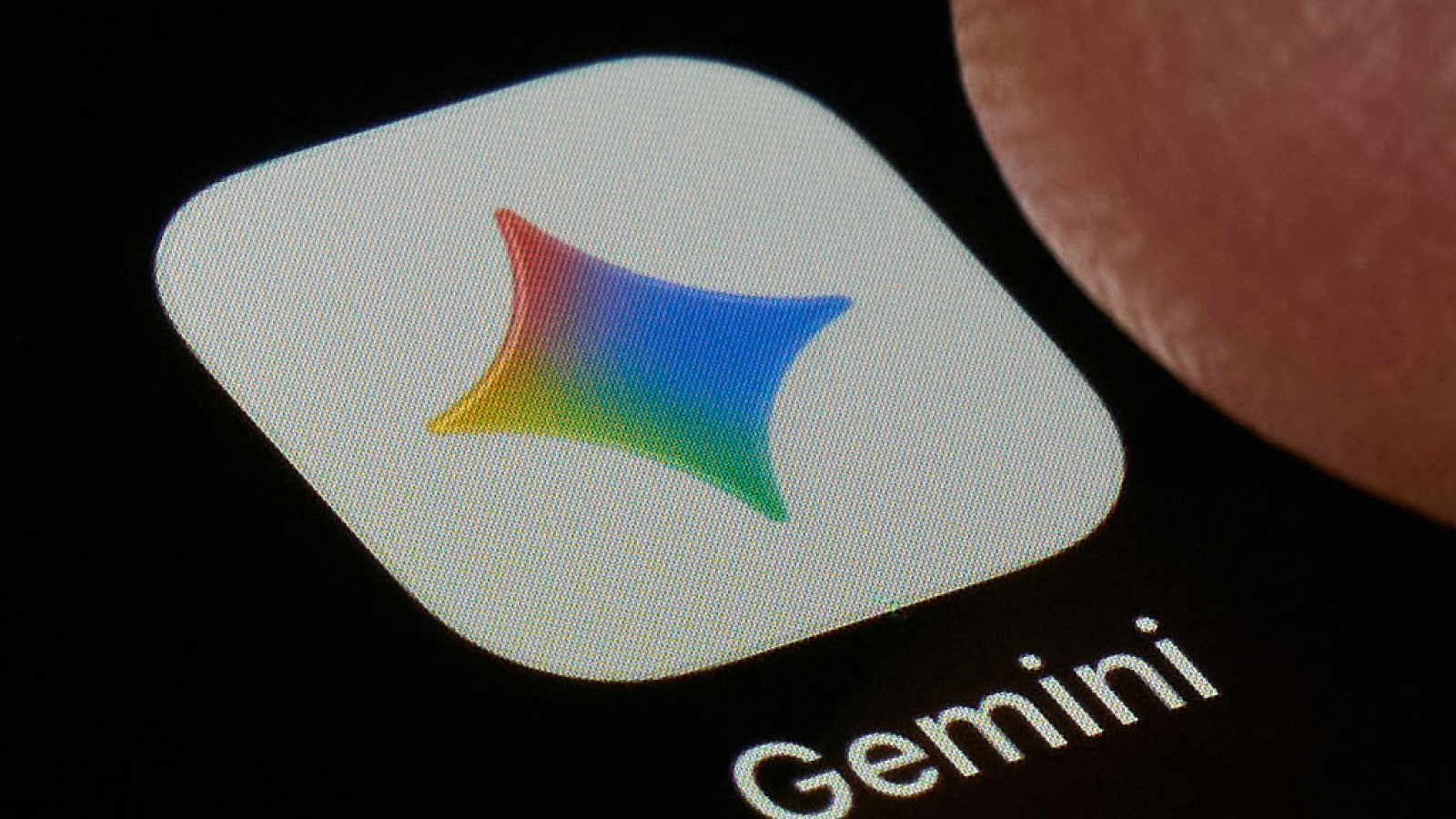 A macro close-up photograph shows the Google Gemini AI application icon on a smartphone screen set against a dark background in San Ferdinando di Puglia, Italy, on May 17, 2026. The icon displays a colorful four-pointed star with a gradient ranging from red and orange through blue and green, positioned on a white rounded-rectangle background. The Gemini wordmark appears in white below the icon. A fingertip is partially visible at the upper right. Gemini is Google's flagship generative AI assistant, representing a major competitor in the consumer-facing large language model market as of May 2026. (Photo Illustration by Matteo Della Torre/NurPhoto via Getty Images)
